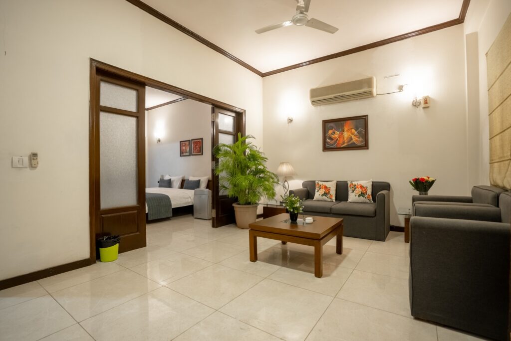 Spacious serviced apartment living room in Gurgaon with sofa seating, coffee table, indoor plants, and an adjoining bedroom